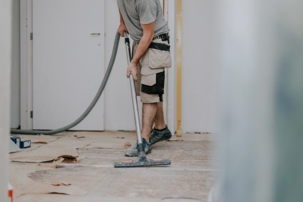 A young caucasian brunette man unrecognizable in work clothes on the left is vacuuming the floor with a construction vacuum cleaner in a room in an old house where renovations are underway, close-up view from the bottom with selective focus.Construction work concept.