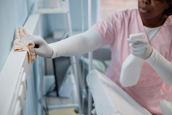 Black person cleaning hospital room with disinfectant spray and cloth, ensuring hygienic environment for patients and staff, while focusing on keeping surfaces sanitized